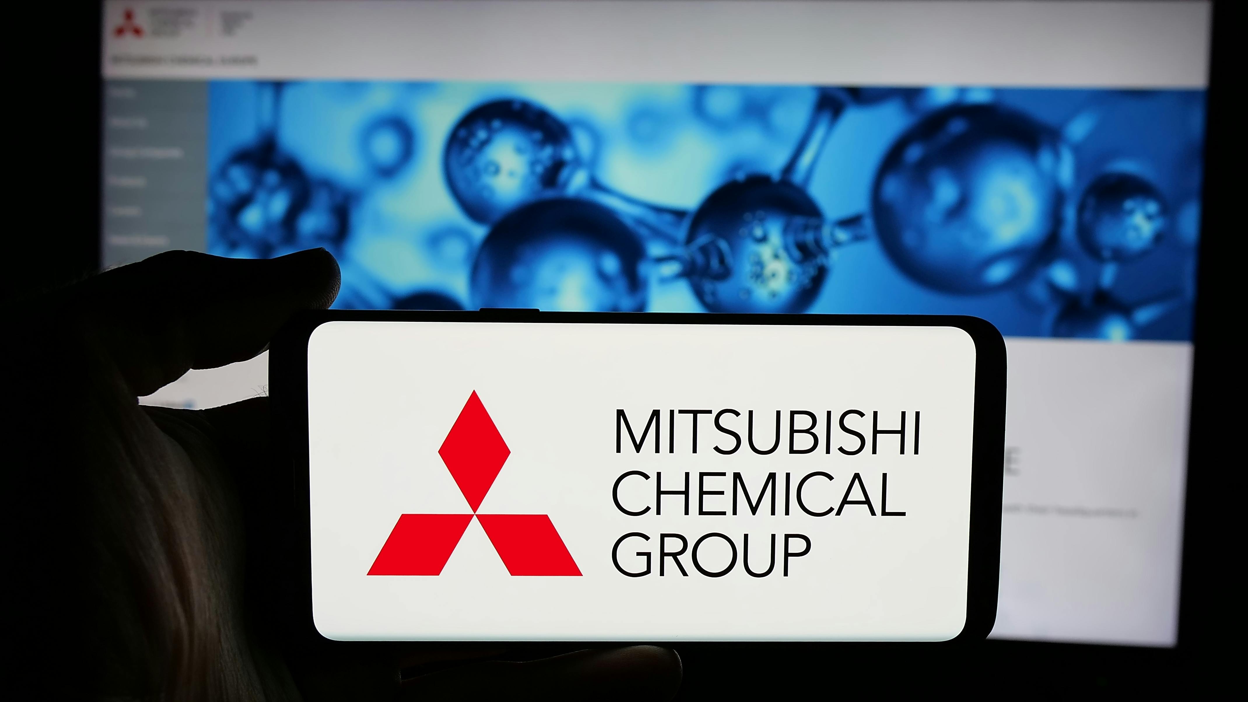 Person holding cellphone with logo of company Mitsubishi Chemical Group Corporation on screen in front of business webpage. Focus on phone display.