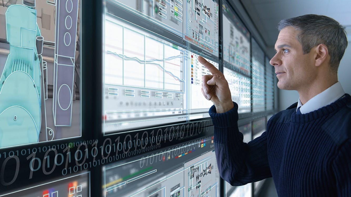 man stands in front of wall of computer screens and selects components on touchscreen