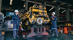 2 men in work overalls and hard hats stand beside large diesel engine
