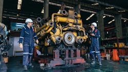 2 men in work overalls and hard hats stand beside large diesel engine 2 men in work overalls and hard hats stand beside large diesel engine