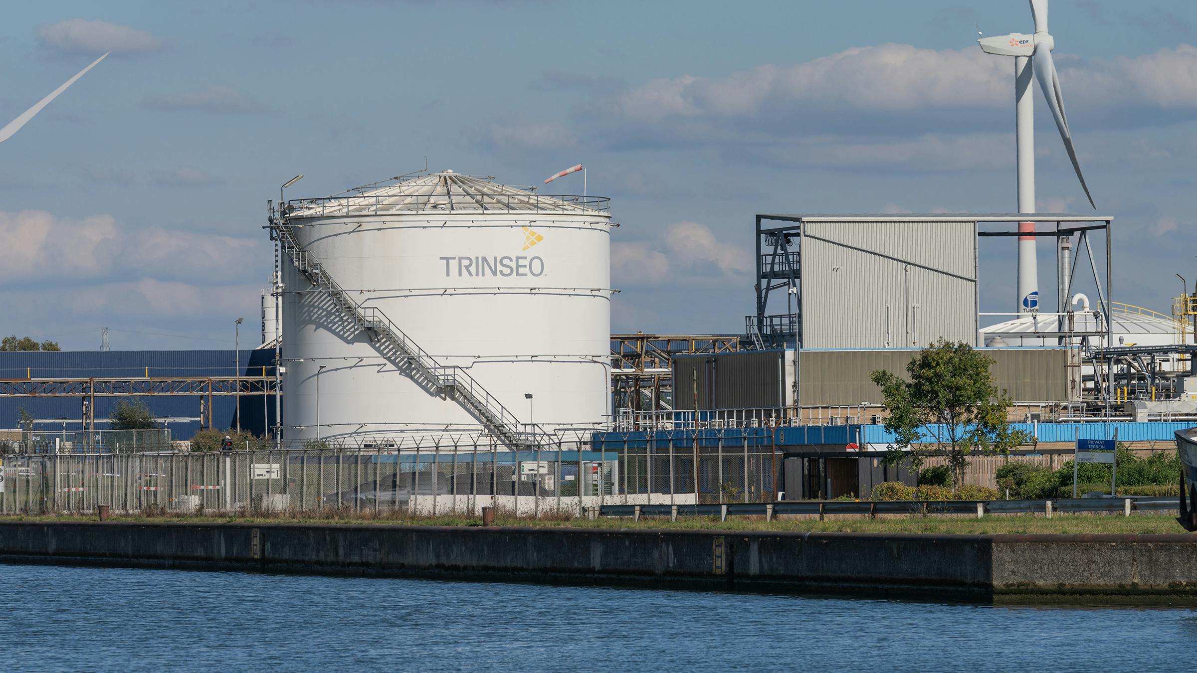 Industrial view of the Trinseo chemical plant with storage tank, wind turbines, and canal water in the foreground under a partly cloudy blue sky. Tessenderlo-Ham, Belgium