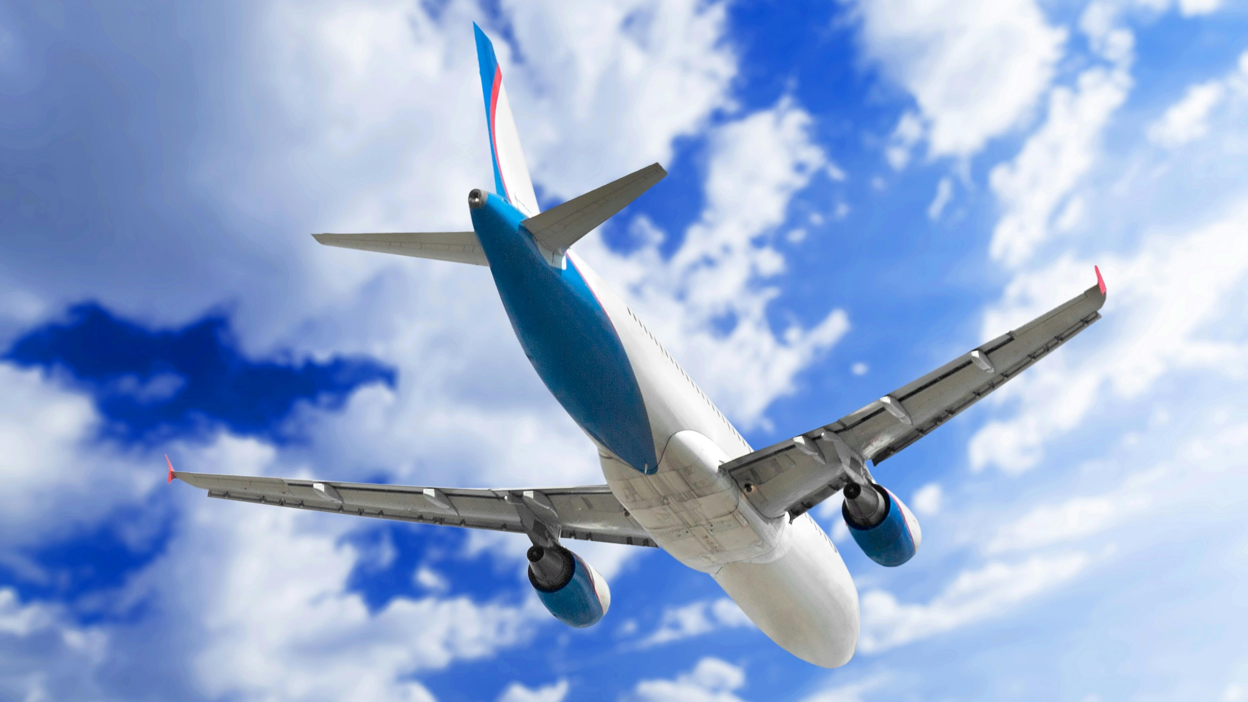 Commercial passenger aircraft photographed from below during flight, showing its white fuselage with blue tail fin and wing accents against a blue sky with white clouds.