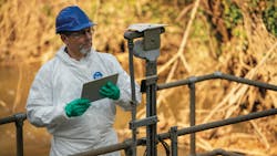 Industrial worker wearing personal protective equipment including a blue hard hat, safety glasses, white Tyvek protective coverall, and bright green chemical-resistant gloves, holding a tablet or clipboard while standing next to industrial piping and equipment. Industrial worker wearing personal protective equipment including a blue hard hat, safety glasses, white Tyvek protective coverall, and bright green chemical-resistant gloves, holding a tablet or clipboard while standing next to industrial piping and equipment.