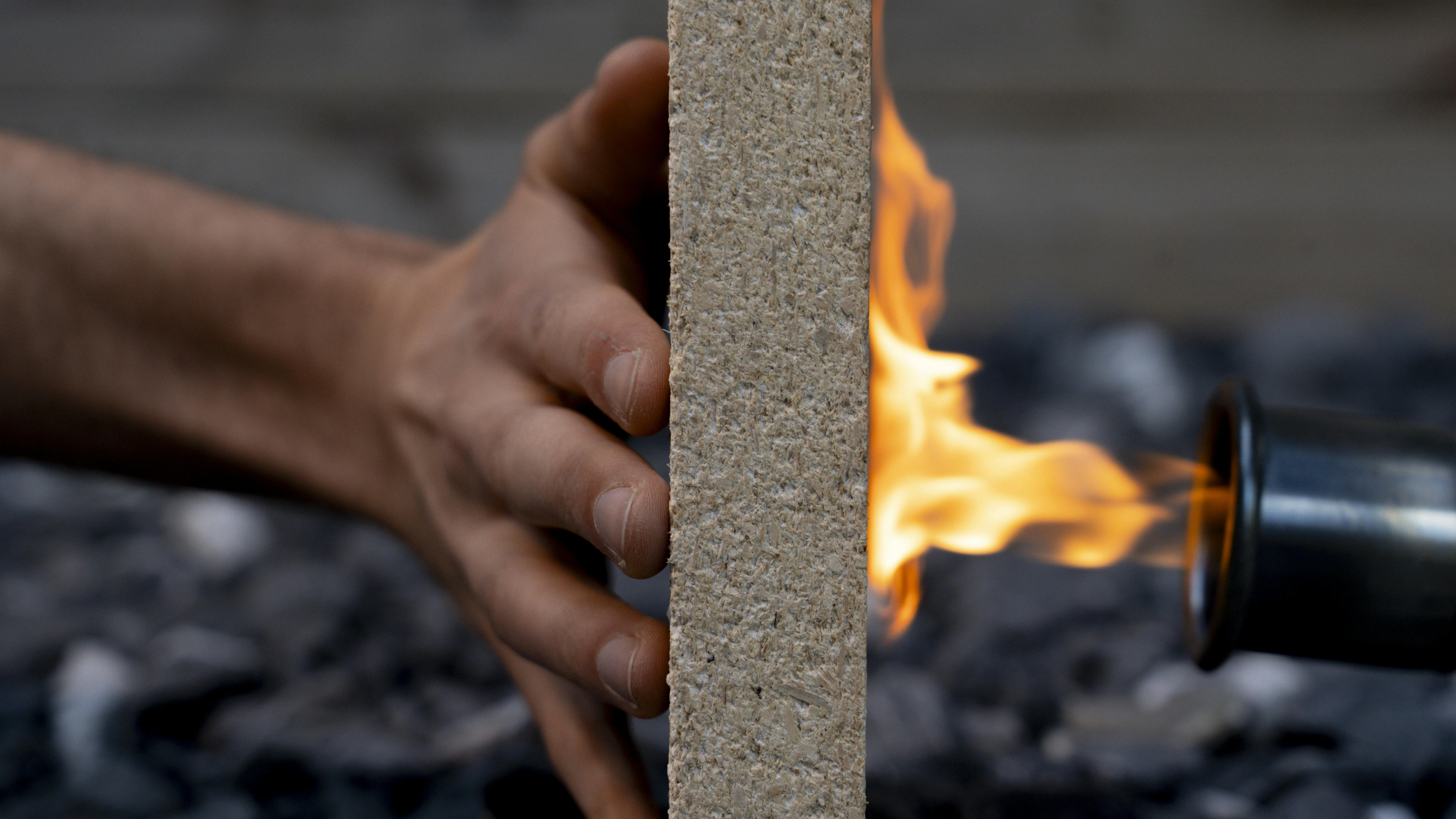 A hand holds a composite board upright while a blowtorch flame burns the opposite side, demonstrating fire resistance.