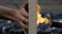 A hand holds a composite board upright while a blowtorch flame burns the opposite side, demonstrating fire resistance.
