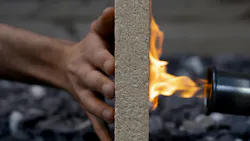 A hand holds a composite board upright while a blowtorch flame burns the opposite side, demonstrating fire resistance. A hand holds a composite board upright while a blowtorch flame burns the opposite side, demonstrating fire resistance.