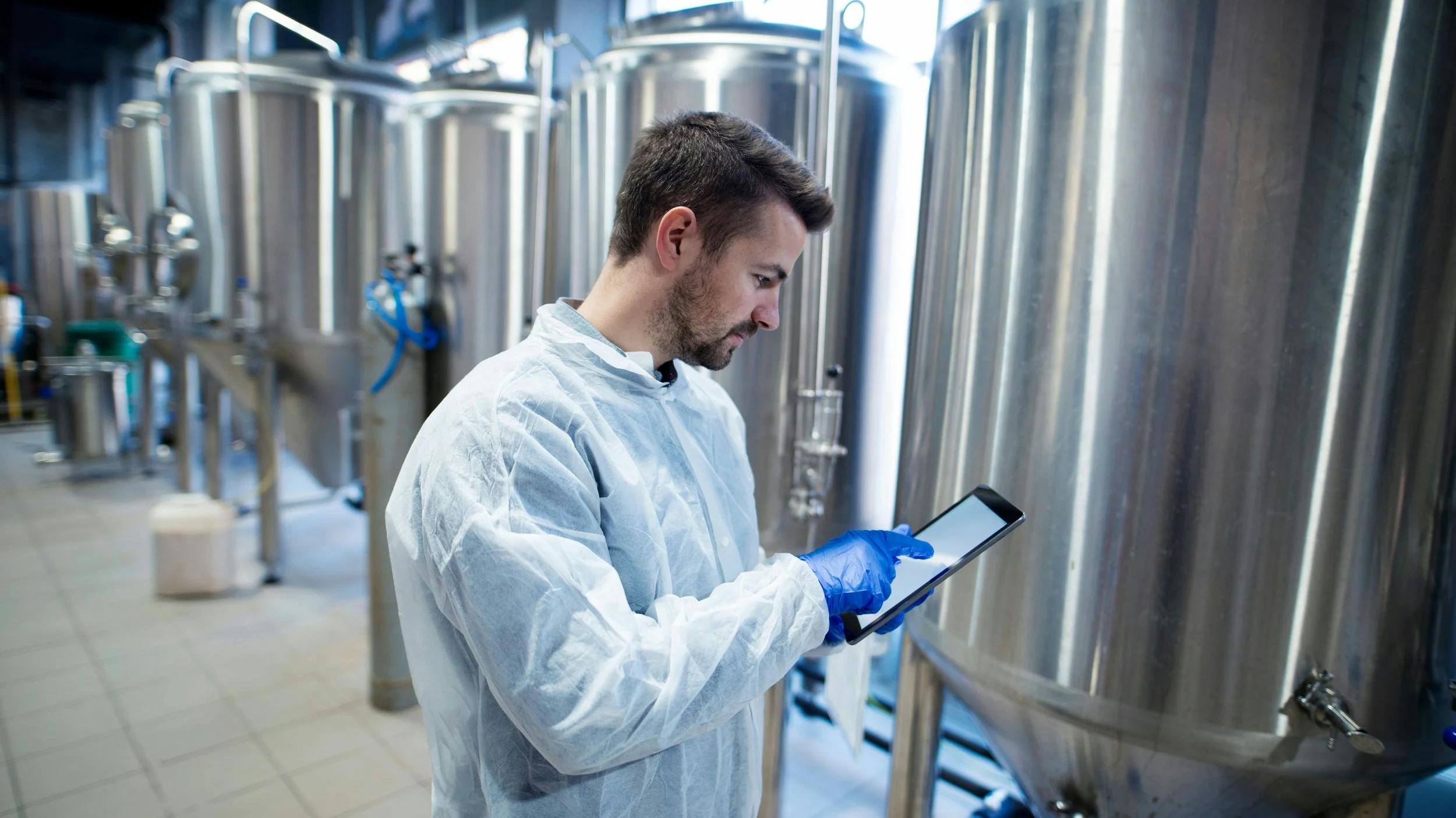 A technician in a white protective coat uses a tablet beside large stainless steel industrial tanks.