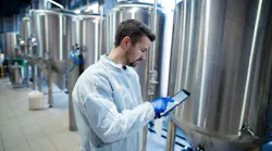 A technician in a white protective coat uses a tablet beside large stainless steel industrial tanks.