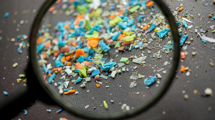 A magnifying glass examining colorful microplastic particles scattered on a dark surface.