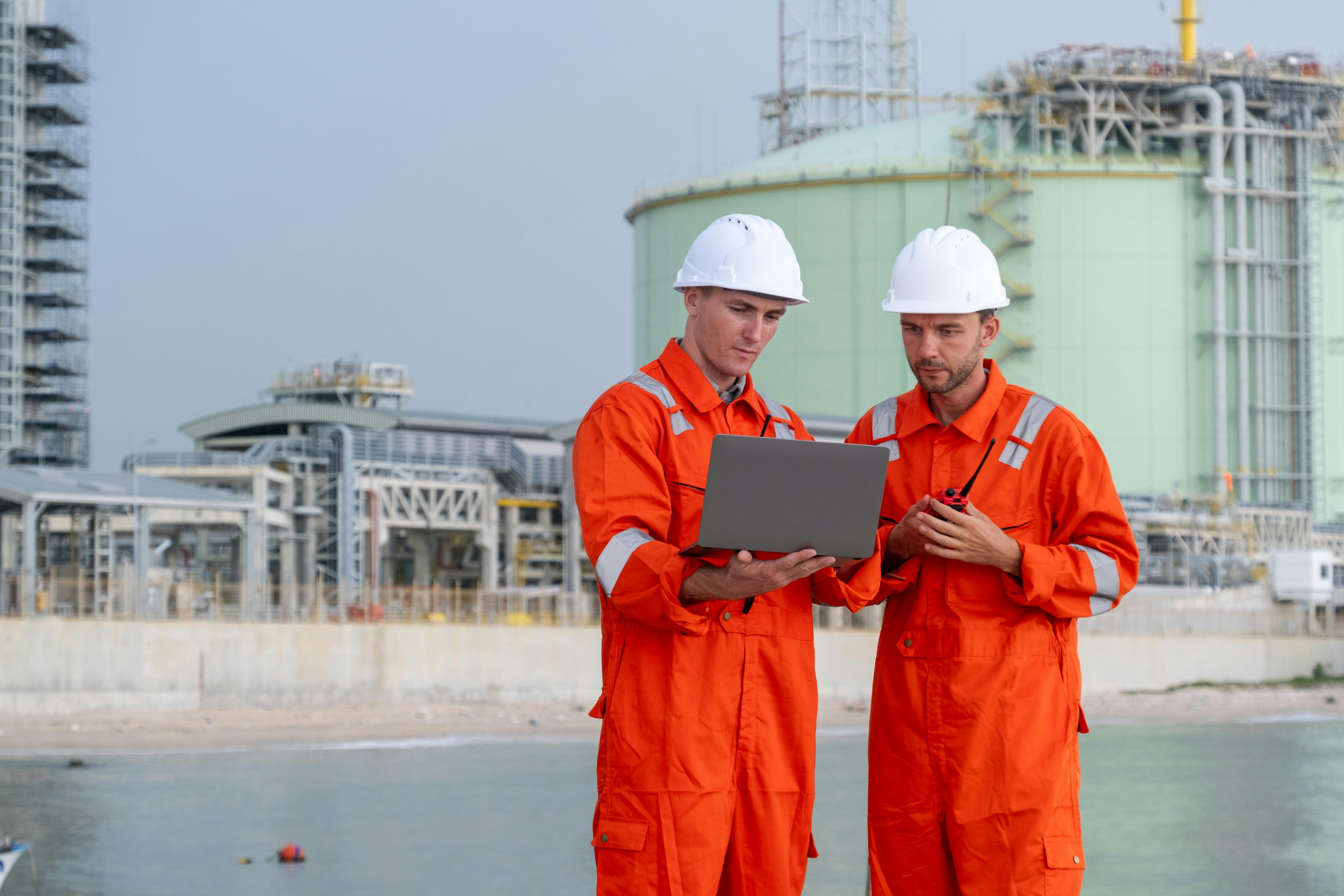 Two engineers in orange coveralls and hard hats review a laptop at an industrial waterfront facility.