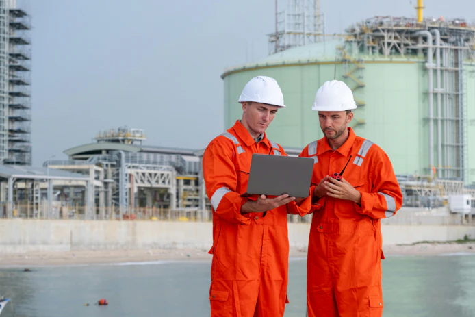 Two engineers in orange coveralls and hard hats review a laptop at an industrial waterfront facility.