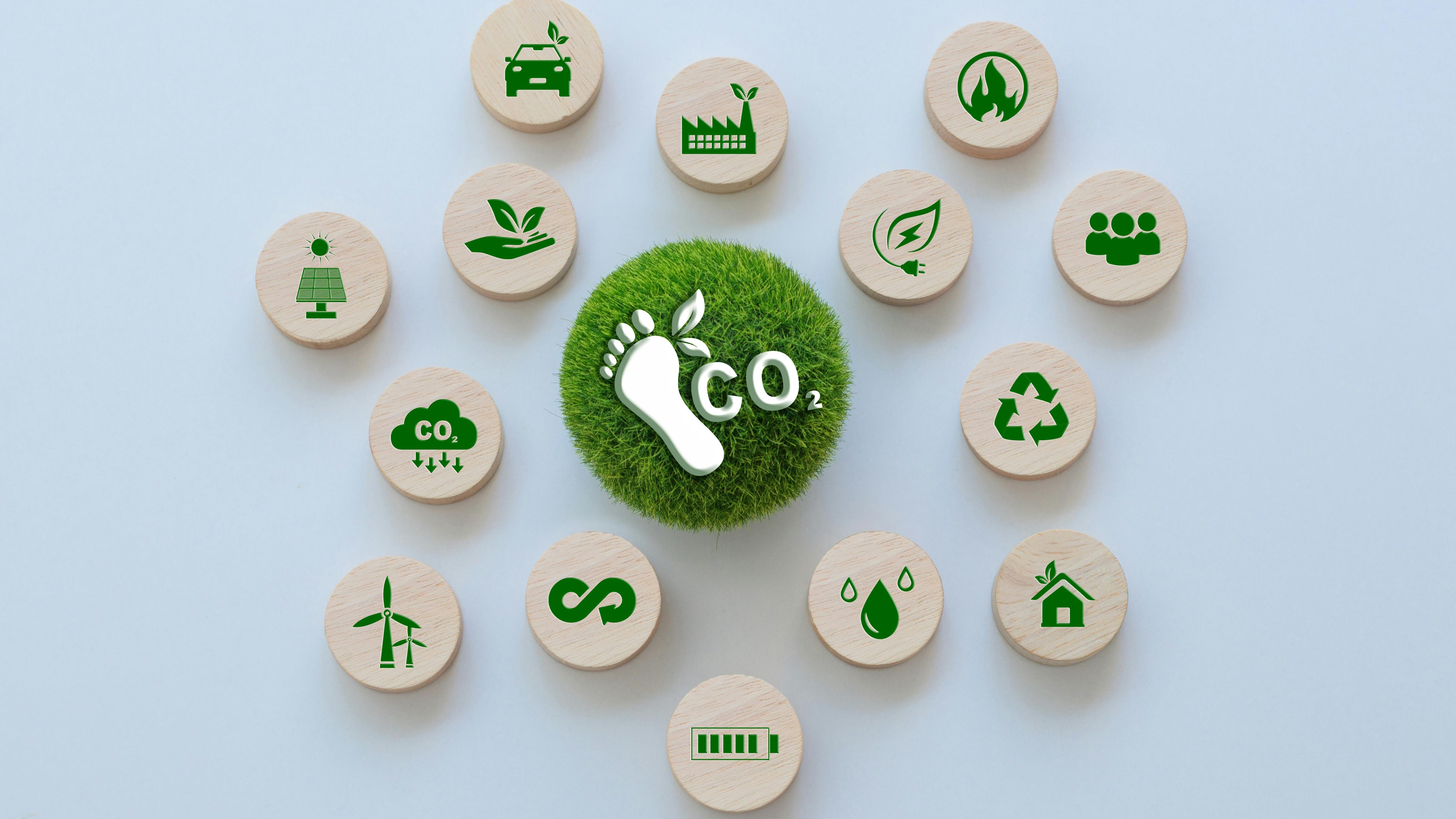Circular arrangement of sustainability and clean-energy icons surrounding a grass-covered sphere with a carbon footprint and CO2 symbol, representing carbon reduction, renewable energy, recycling and environmental responsibility.