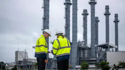 Two engineers in high-visibility jackets and hard hats review documents at an industrial plant