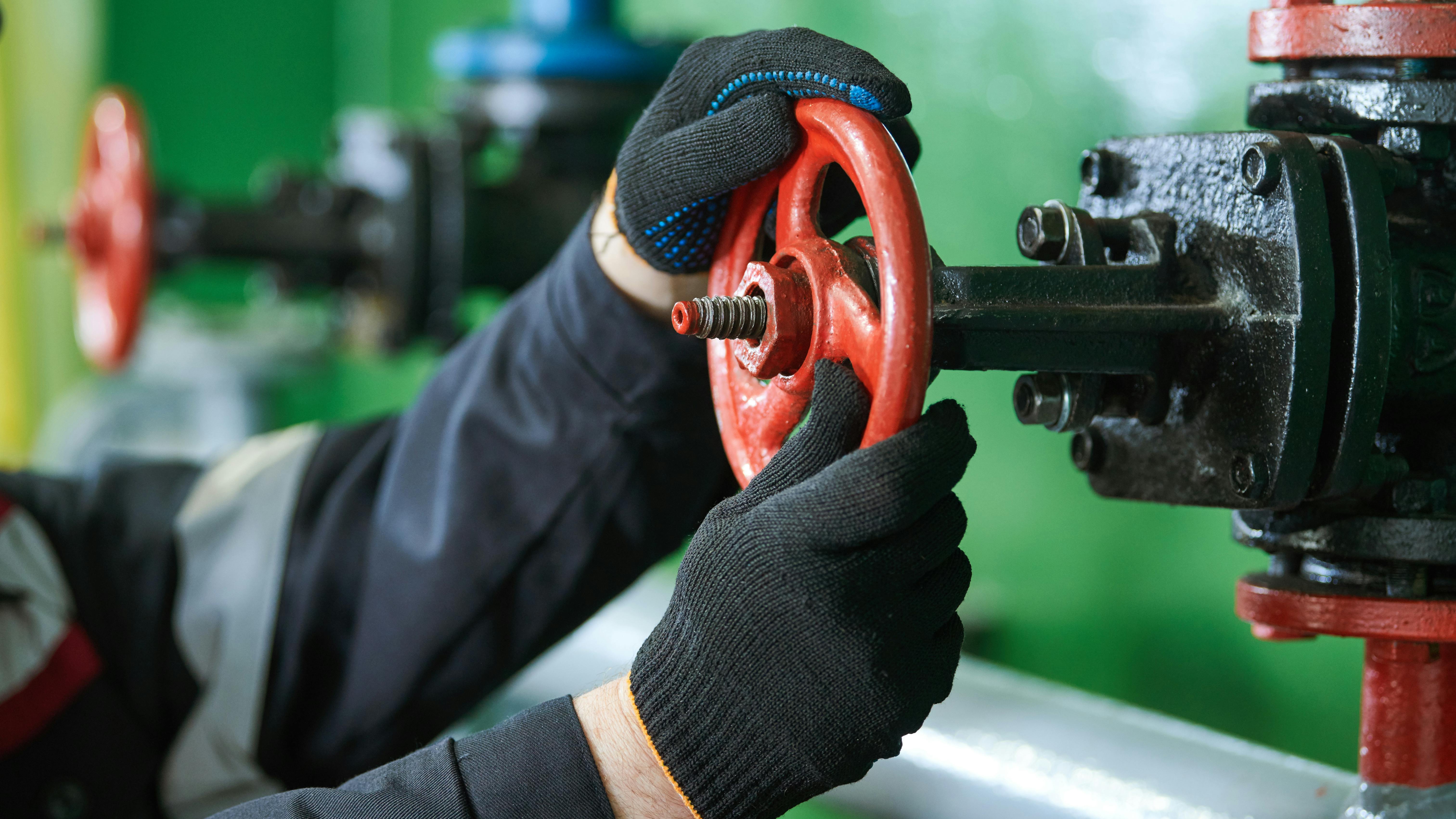 A gloved worker manually operates a red handwheel gate valve on an industrial pipeline.