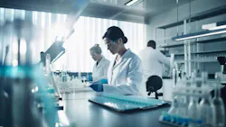 Scientists in white lab coats and safety glasses work at a bright, modern laboratory bench with glassware.