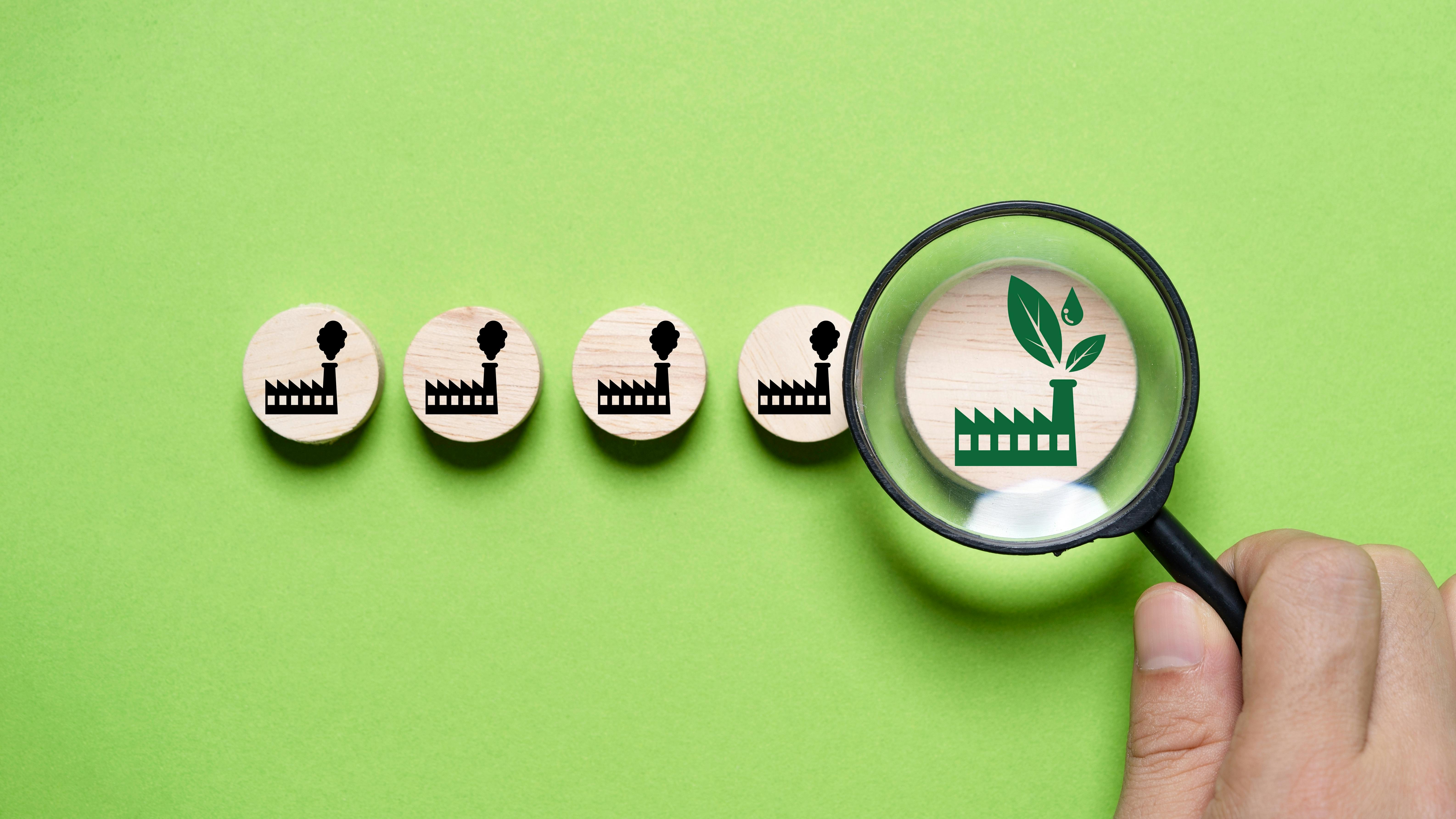A magnifying glass highlights a green eco-friendly factory icon among polluting factory icons on wooden discs.