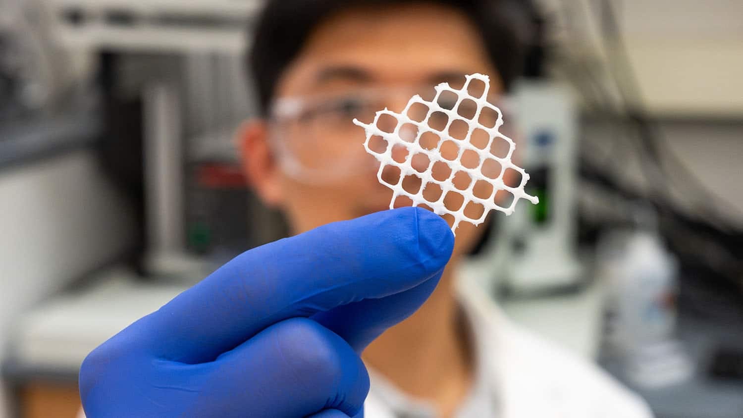 a researcher in a white labcoat and purple gloves holds a piece of white mesh up in front of the camera