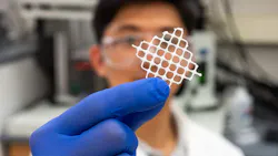 a researcher in a white labcoat and purple gloves holds a piece of white mesh up in front of the camera