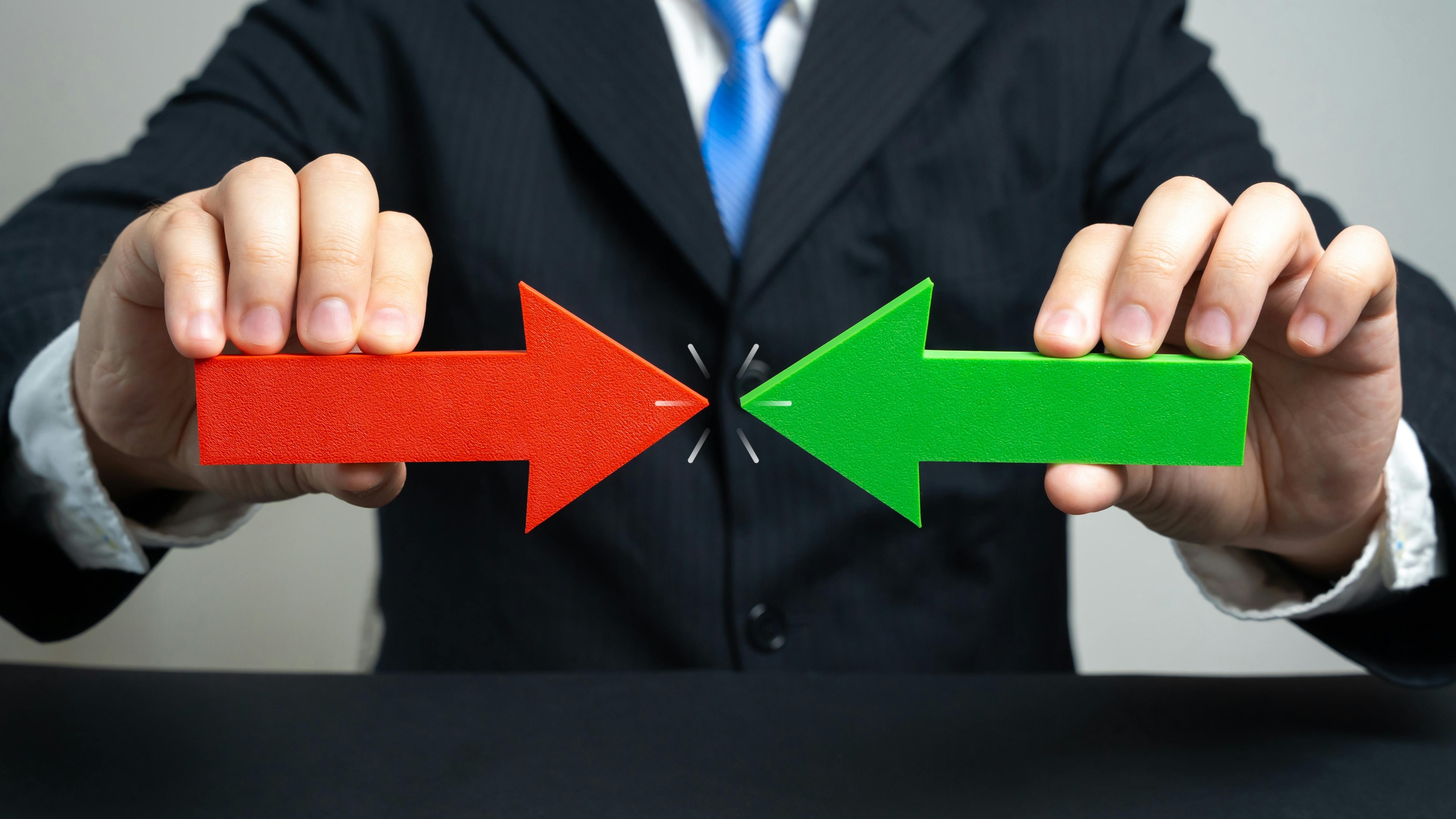 Person in a business suit holds a red arrow and a green arrow pointing toward each other, symbolizing negotiation, alignment, or convergence.