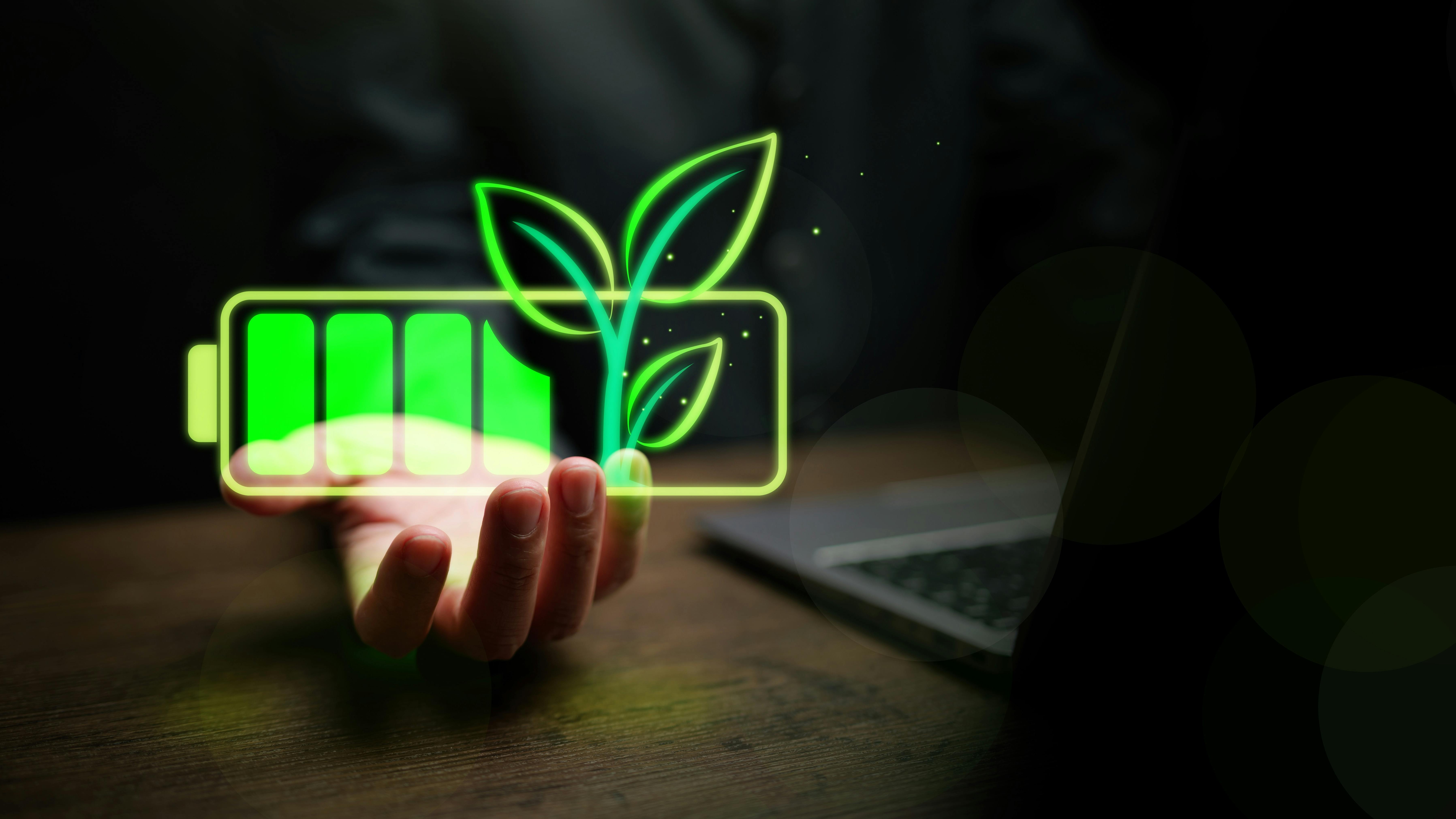 Glowing green battery icon with sprouting leaves held above a desk with laptop computer