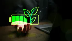 Glowing green battery icon with sprouting leaves held above a desk with laptop computer