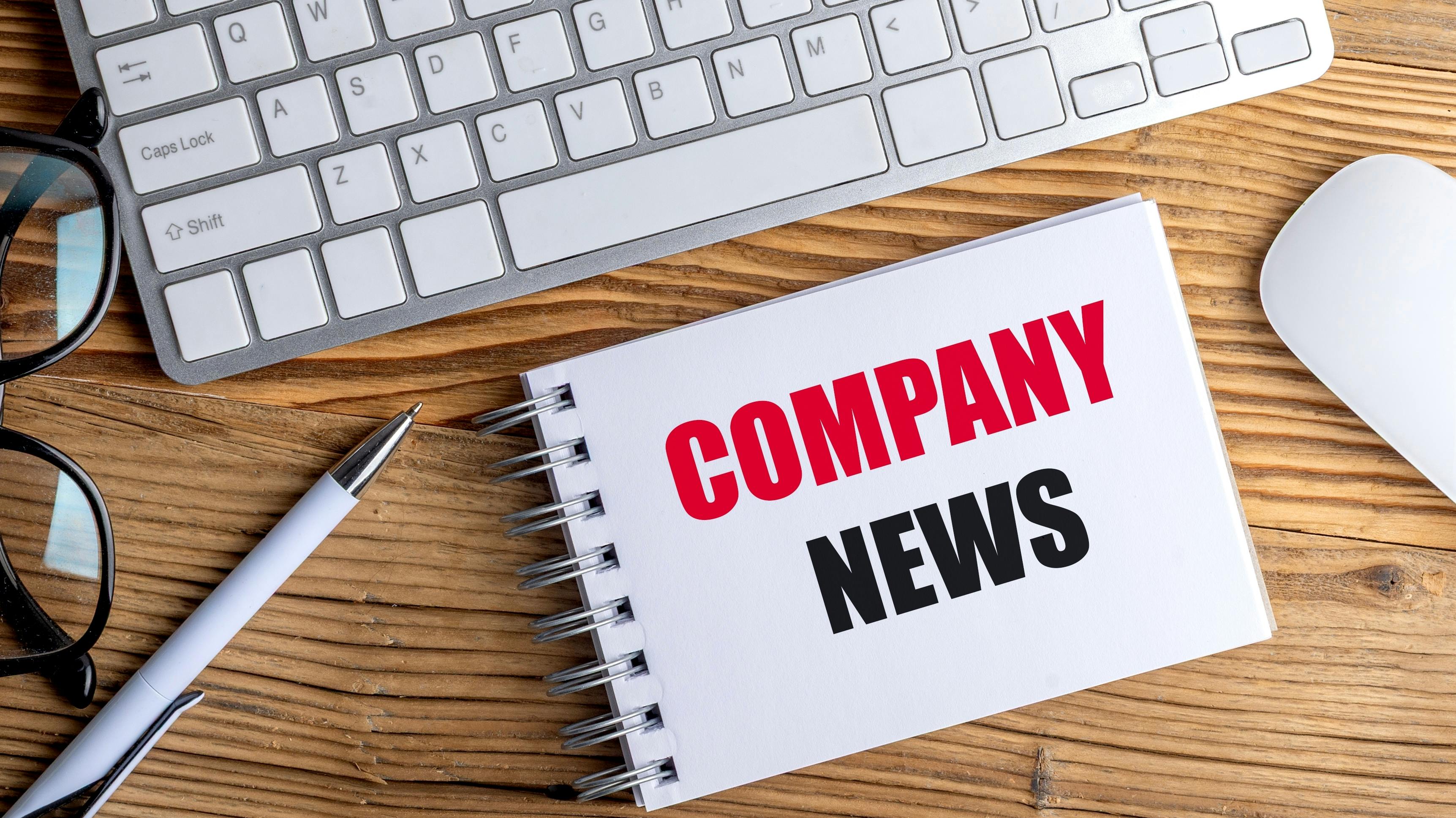 Notepad with text company news on a wooden desk with keyboard, mouse, pen and glasses .